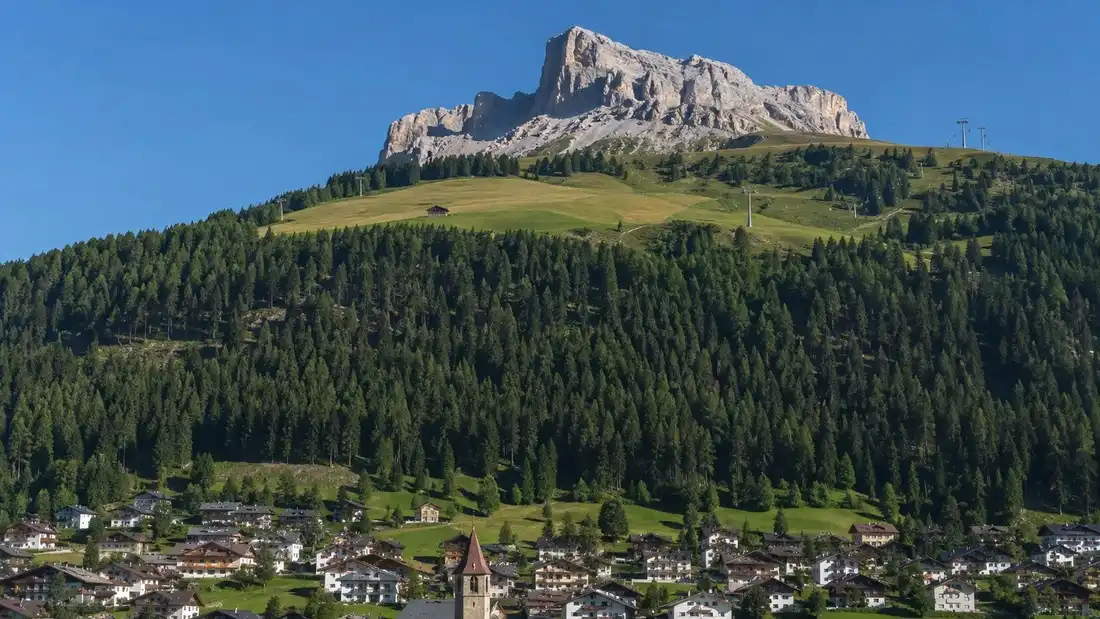 Zalesněný svah Haunoldu nad Innichenem s horskou loukou, chatou a dolomitskými skalami nad ní